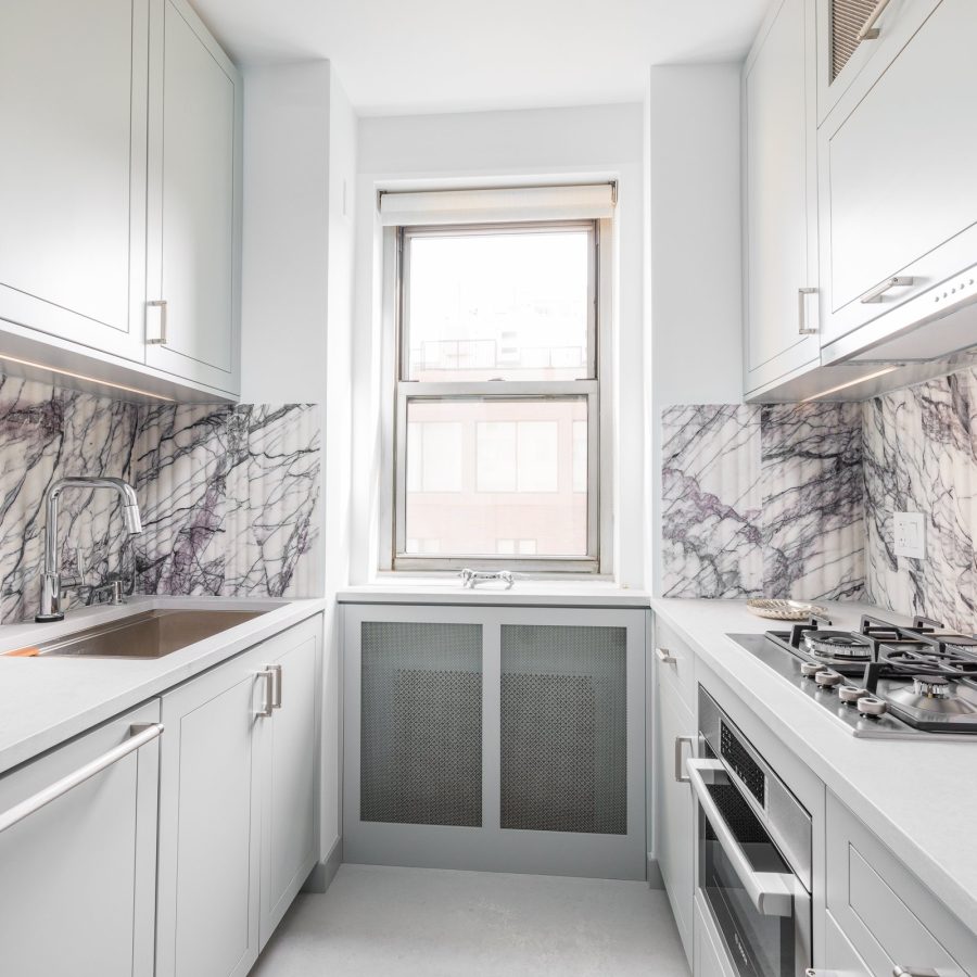 10389 Kitchen remodeling in Manhattan. Modern galley kitchen with light gray cabinetry, sleek white countertops, and dramatic marble backsplash, featuring a window centered at the far wall.