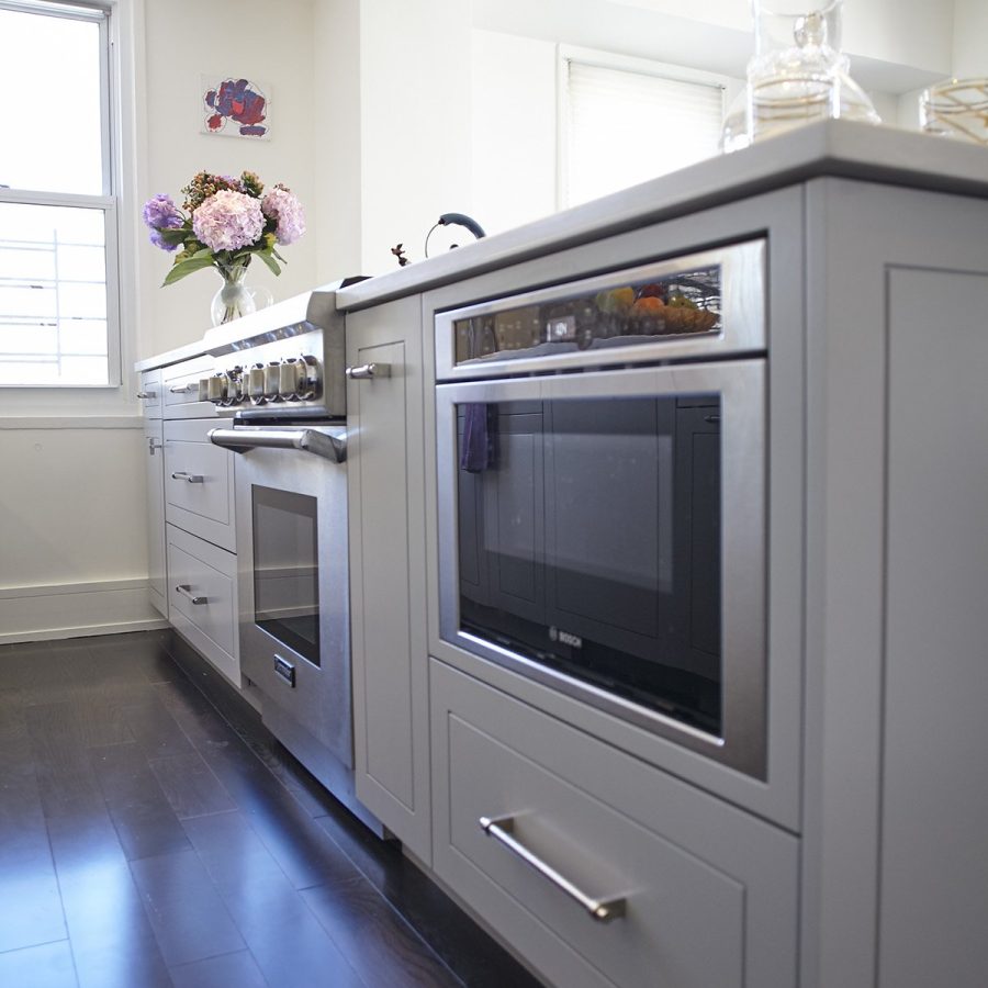 10390 Home remodeling contractor in Lenox Hill. Modern kitchen island with built-in stainless steel oven and microwave, light gray cabinetry, dark hardwood floors, and a vase of fresh flowers on the countertop.
