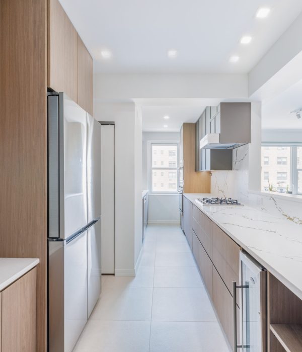 Contemporary galley kitchen featuring sleek flat-panel cabinetry in wood and gray tones, paired with white quartz countertops accented by subtle gold veining. The space includes a built-in gas cooktop, integrated appliances, under-cabinet lighting, and a beverage fridge, with natural light streaming in through a large rear-facing window.