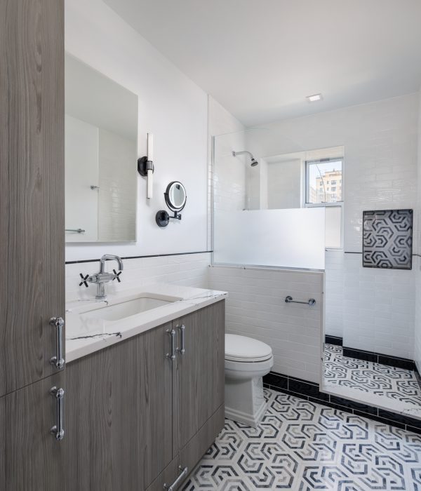Spacious bathroom with a gray floating vanity, large frameless mirror, and black-and-white patterned floor tiles. White walls and a walk-in shower with clear glass doors create a clean, modern aesthetic with subtle vintage accents.