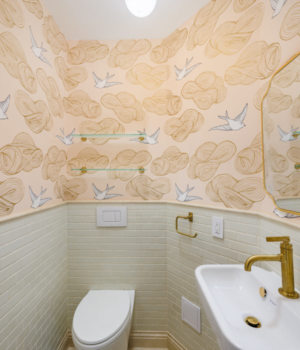 Charming powder room featuring whimsical cloud-and-swallow wallpaper in soft peach tones, complemented by glossy cream subway tiles and hexagon tile flooring. The space includes a wall-mounted toilet, a compact floating sink with gold fixtures, and a sculptural globe light that adds a playful, modern touch.
