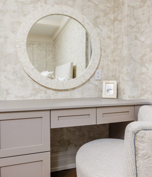 Elegant vanity area featuring a circular mirror framed in stone-like texture, soft beige cabinetry, and a cushioned stool. The design creates a serene, spa-like space for grooming and daily routines, complemented by subtle decorative touches.