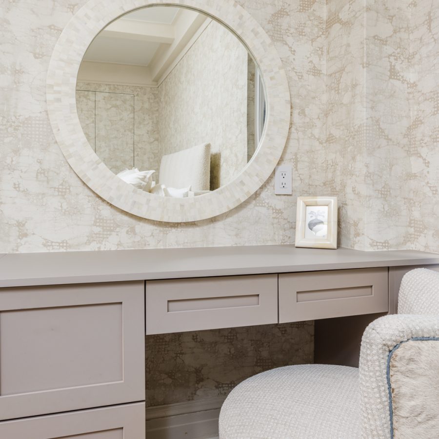 Elegant vanity area featuring a circular mirror framed in stone-like texture, soft beige cabinetry, and a cushioned stool. The design creates a serene, spa-like space for grooming and daily routines, complemented by subtle decorative touches.