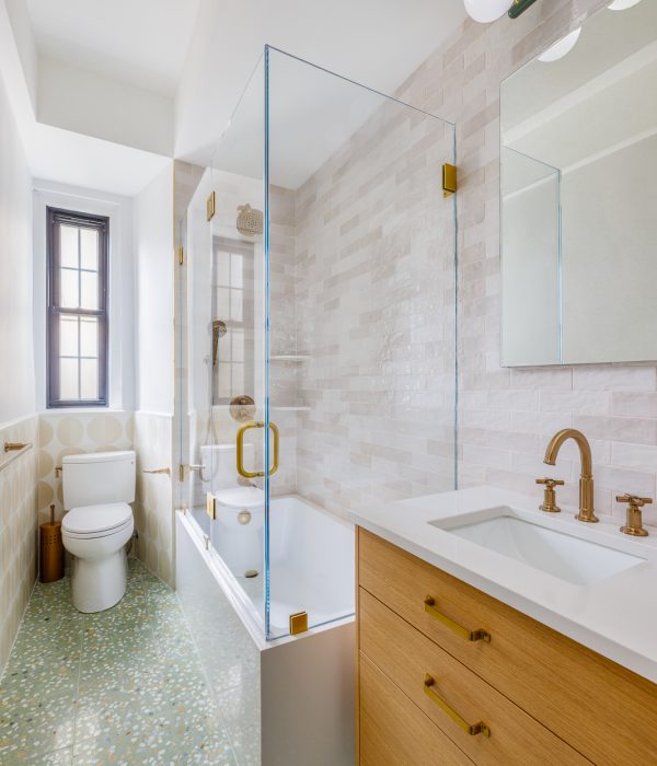 Elegant bathroom featuring a terrazzo-style green floor with multicolored chips, a white soaking tub with frameless glass enclosure and brass hardware, and a warm wood vanity topped with a white quartz sink. The space is finished with cream-toned zellige-style shower tiles, a globe light fixture, and a large window for natural light.