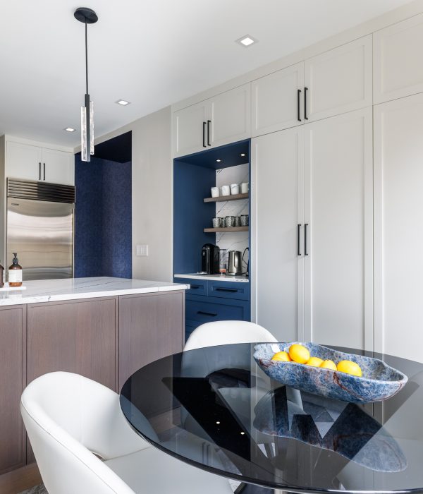 Modern kitchen with a round glass dining table surrounded by white upholstered chairs. The space features wood-accented lower cabinets, marble countertops, and a navy beverage nook with open shelving, creating a cohesive and stylish look.