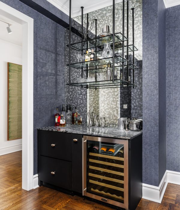 Sophisticated home bar area with deep blue cabinetry, mirrored backsplash, and glass shelving for liquor and glassware. A brass rail detail and built-in wine storage enhance the luxurious design, ideal for entertaining in a compact space.