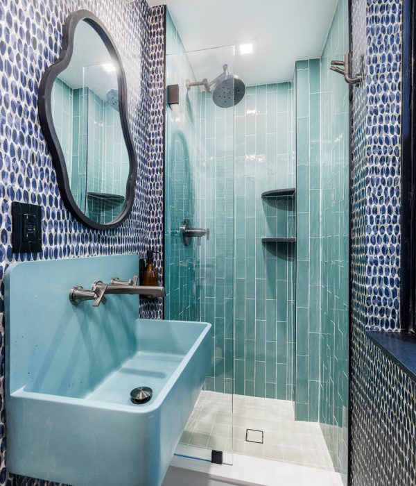 Playful bathroom with patterned blue wall tiles, a light teal vertical-tiled shower, and a matching blue wall-mounted sink. An irregular-shaped mirror adds an eclectic touch, balancing bold color and modern design.