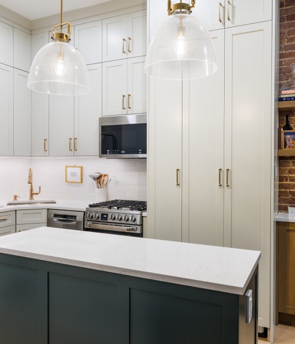 Chic transitional kitchen featuring soft gray shaker cabinets with gold hardware, a deep green island with a white quartz countertop, and a stainless steel appliance suite. A cozy exposed brick coffee nook with open wood shelving adds warmth and character, illuminated by under-cabinet lighting and flanked by globe pendant lights over the island.