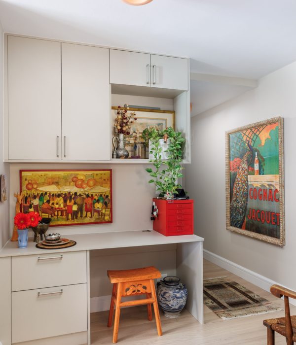 Functional desk nook integrated into white cabinetry with open shelving and vibrant artwork. A colorful painting and potted plant above the workspace add personality and warmth to the otherwise neutral palette, making it ideal for a creative home office corner.
