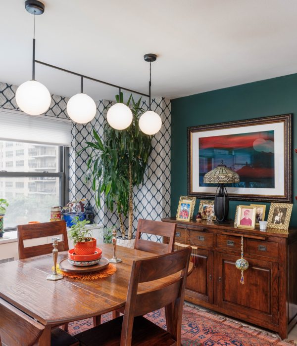 Bright dining room with mid-century style wooden furniture and a contemporary light fixture featuring globe-shaped bulbs. The walls are painted a deep green and accented with bold artwork, plants, and patterned curtains, blending modern and eclectic design elements.