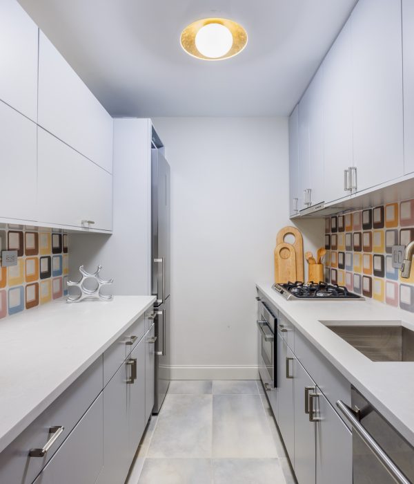 Bright, narrow kitchen with all-white cabinetry contrasted by a geometric patterned backsplash in warm earth tones. Streamlined appliances and marble-like flooring enhance the minimalist yet playful design.