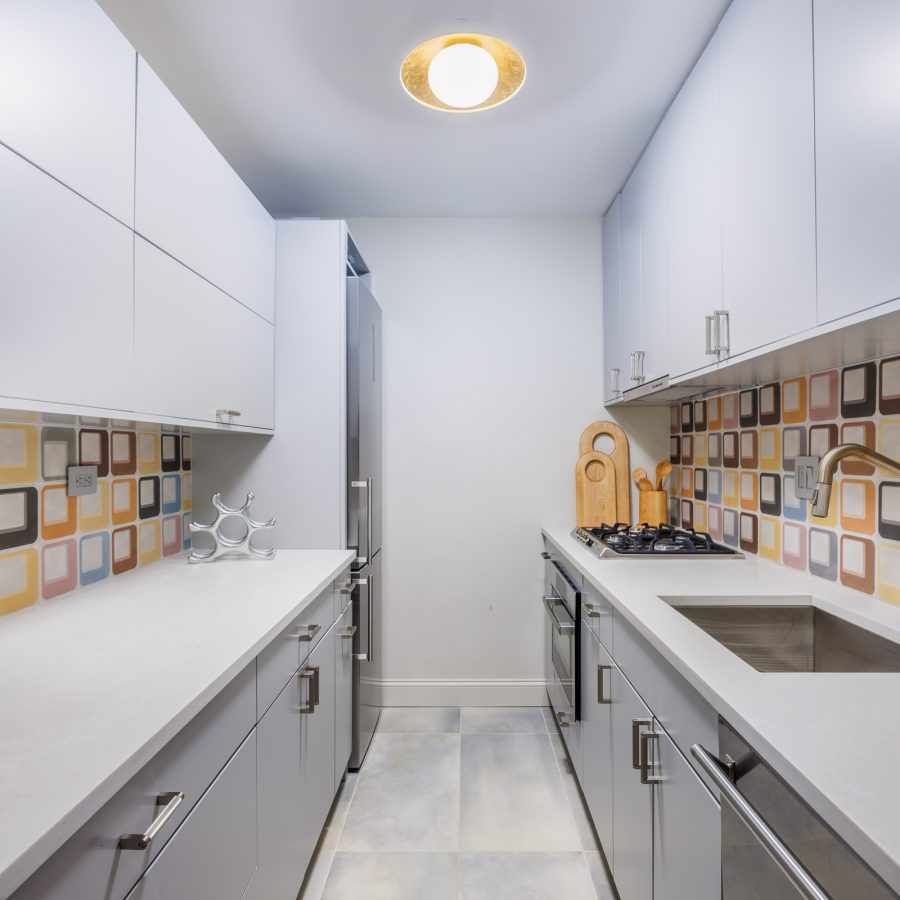 Bright, narrow kitchen with all-white cabinetry contrasted by a geometric patterned backsplash in warm earth tones. Streamlined appliances and marble-like flooring enhance the minimalist yet playful design.