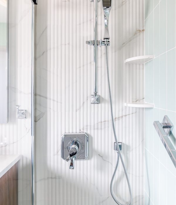 Detail view of a shower with slim vertical white tiles and chrome fixtures, including a rain showerhead and handheld sprayer. Built-in shelving adds practical storage in this clean, minimalist bathroom design.