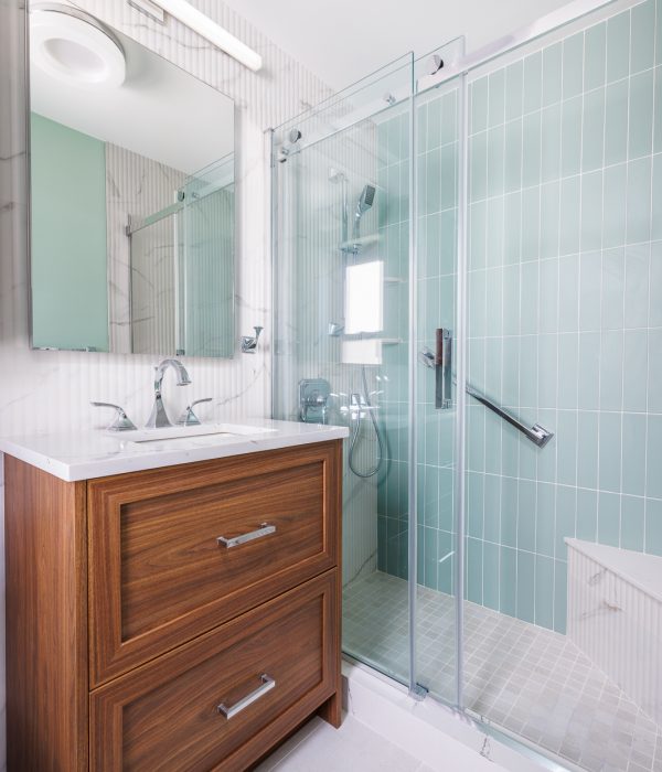 Bright bathroom with a medium-tone wood vanity and white countertop, complemented by a frameless glass shower with green vertical tiles. The design blends warm natural finishes with fresh modern materials.