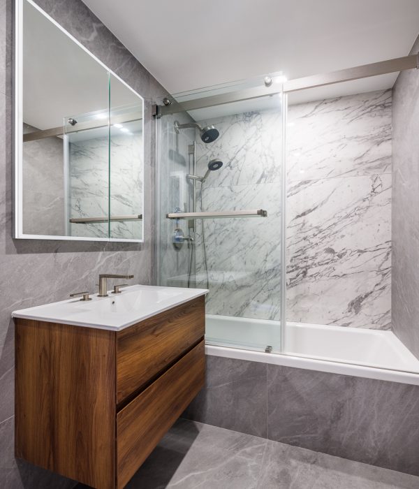 Elegant bathroom featuring a wood floating vanity with a white countertop, paired with gray marble walls surrounding a tub-shower combo. A frameless mirror and soft lighting add a refined, spa-like atmosphere.