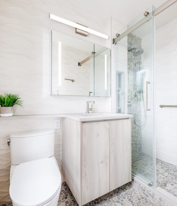 Bright, spa-inspired bathroom featuring a floating light wood vanity with a white quartz countertop, a backlit mirror, and a frameless glass shower enclosure. The space is unified by river rock pebble flooring that extends into the shower, paired with soft white textured wall tiles and brushed nickel fixtures for a calming, organic aesthetic.