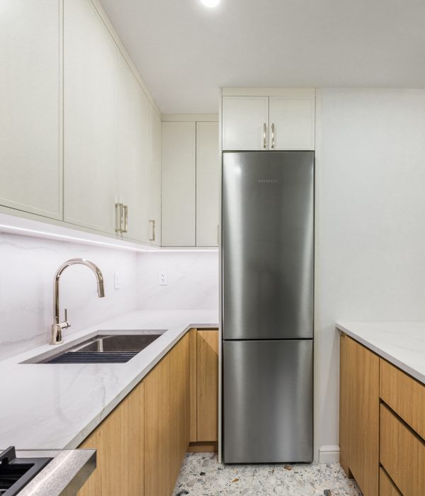 Compact modern kitchen with two-tone cabinetry, featuring white upper cabinets and warm wood lower drawers, accented by white quartz countertops and backsplash. The space includes a sleek stainless steel refrigerator, under-cabinet lighting, and terrazzo tile flooring for a contemporary touch with visual texture.