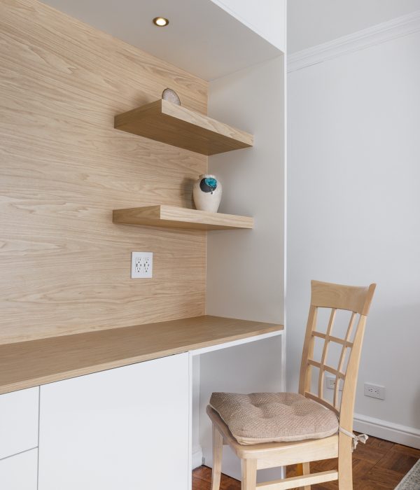 Minimalist home office nook with a built-in desk featuring light wood surfaces and white cabinetry. Floating wooden shelves hold simple decorative accents, while recessed lighting above adds a warm glow. A matching wooden chair with a cushioned seat completes the compact, functional workspace set against clean white walls and hardwood flooring.