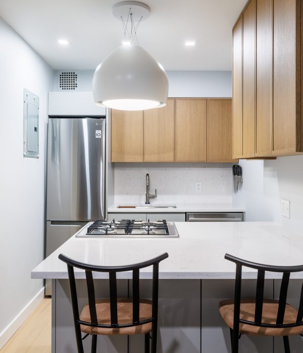 Compact modern kitchen with white countertops and backsplash, complemented by wood cabinetry and a stainless steel refrigerator. A central peninsula with a built-in cooktop serves as a casual dining area with two black spindle-back chairs, illuminated by a large white pendant light for a clean, contemporary feel.
