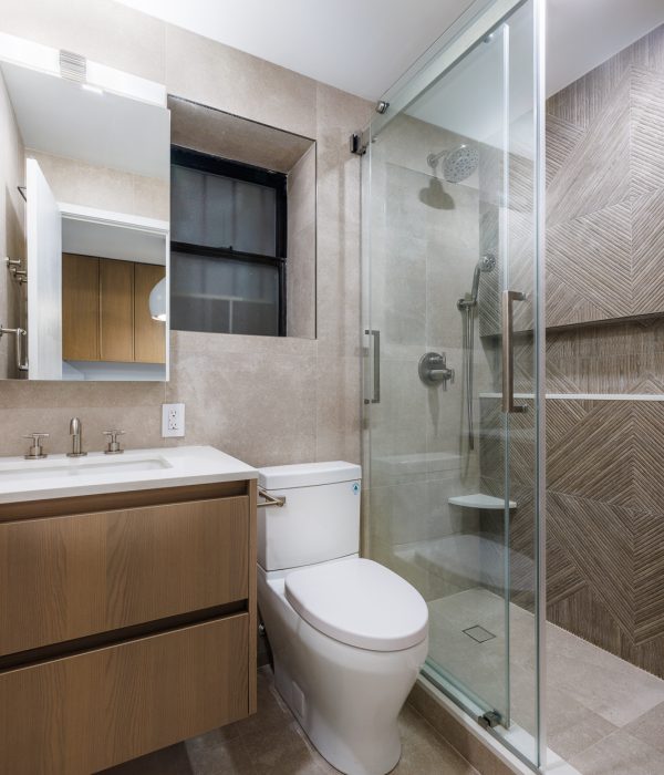 Compact bathroom featuring a wood vanity with a white countertop, paired with a frameless glass shower enclosed by tan herringbone wall tiles. A black-framed window adds contrast and natural light to the warm, contemporary design.