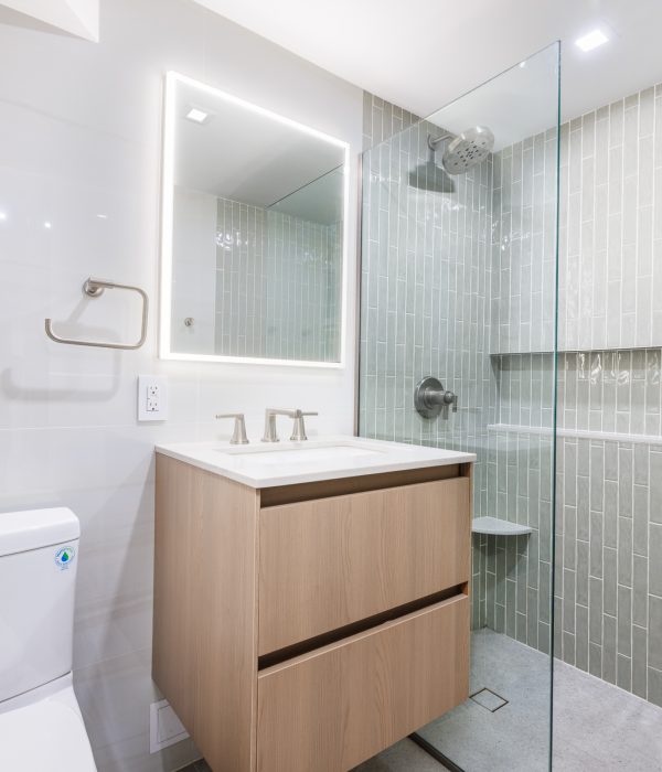 Modern bathroom with a backlit rectangular mirror, light wood floating vanity, and a frameless glass shower enclosure. The shower features vertical mint-green tiles, chrome fixtures, and minimalist white walls for a fresh, airy look.