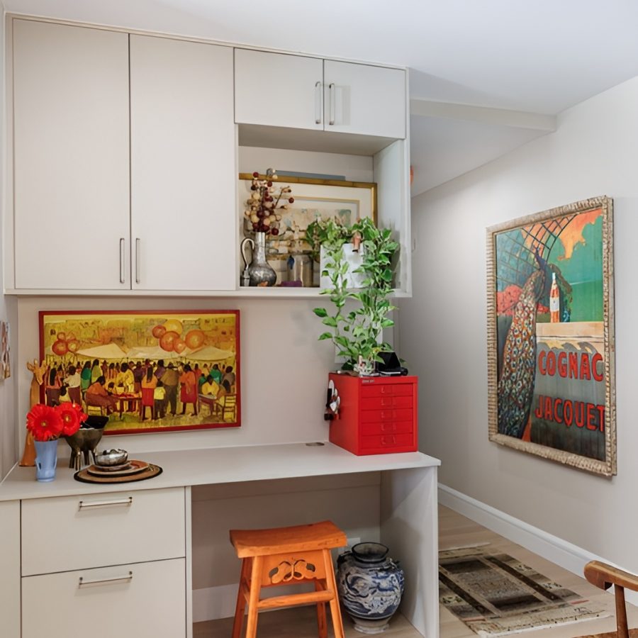 Warm Contemporary Manhattan Desk Area Bright and colorful home office nook with sleek white cabinetry, light wood flooring, and vibrant decorative accents. A built-in desk area features a small orange stool, red storage drawers, and potted plants. Framed artwork with bold colors lines the wall above the desk and in the hallway, including a large vintage-style "Cognac Jacquet" poster. Open shelving displays ceramics, vases, and framed art, while a traditional blue-and-white porcelain jar sits below the desk. The space blends contemporary cabinetry with eclectic, artistic charm.