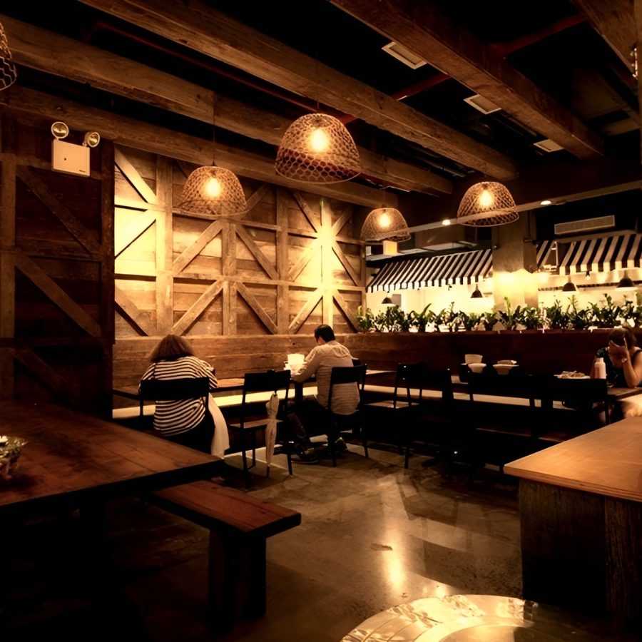 Screenshot 2025-07-24 141501 (1) Cozy rustic restaurant interior featuring reclaimed wood paneling, exposed ceiling beams, and warm ambient lighting from woven pendant lamps. The space includes long communal tables, simple black chairs, and a semi-open kitchen with a striped awning detail. The earthy tones and intimate atmosphere create a relaxed dining experience with a touch of vintage charm. Commercial interior design and remodeling in Manhattan.