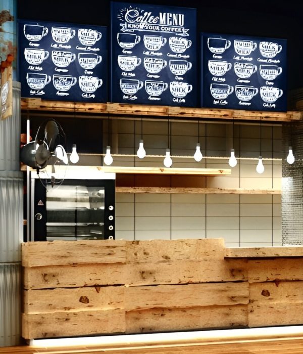 Industrial coffee shop interior with rustic wood paneling, corrugated metal accents, and exposed structural columns featuring distressed paint. The counter area includes hanging pendant lights, open shelving, and a large chalkboard-style menu with hand-drawn coffee illustrations. The combination of raw textures and warm wood tones creates a trendy, urban vibe suited for a casual café environment.
