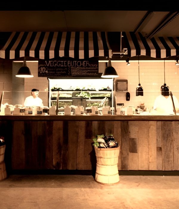 Casual café counter with a rustic wood front, black-and-white striped awnings, and white subway tile walls. Pendant lights hang above the serving area, where staff prepare fresh greens and salads displayed behind the counter. A customer stands at the counter while baskets of chips and a refrigerated display case add to the approachable, market-inspired ambiance.