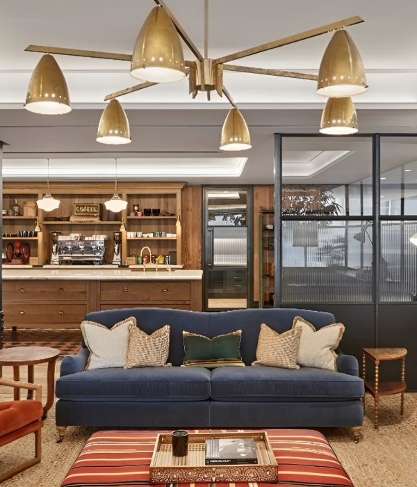Stylish lounge area featuring a navy blue sofa with patterned throw pillows, a burnt orange accent chair, and a striped ottoman serving as a coffee table. A brass mid-century chandelier hangs overhead, illuminating the warm wood finishes and industrial glass partitions that frame the space. The backdrop includes an inviting coffee bar with open shelving, pendant lights, and vintage-inspired details, blending comfort with modern sophistication.