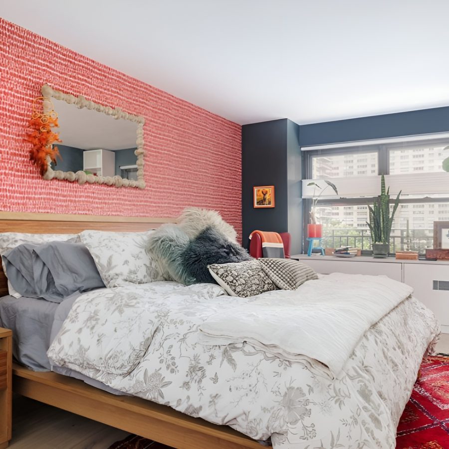 Bold Boho Manhattan Bedroom Eclectic bedroom featuring a bold red patterned accent wall behind a wooden bed frame dressed in floral bedding with layered textured pillows. A mix of vintage and modern décor includes a mid-century lamp, indoor plants, and built-in shelving with colorful accents. A large window brings natural light to the space, complemented by a red patterned rug for added warmth and character. Complete home remodeling in Manhattan.