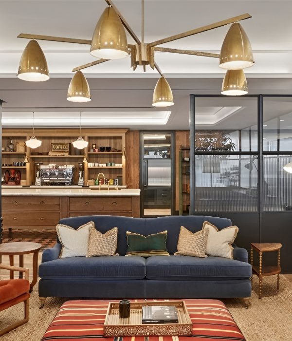 A stylish, modern lounge with a mid-century aesthetic featuring a navy blue velvet sofa, accent pillows, and a burnt orange armchair, set atop a woven natural rug. A large brass chandelier hangs overhead, while the background includes a cozy coffee station framed in rustic wood cabinetry and fluted glass office partitions.