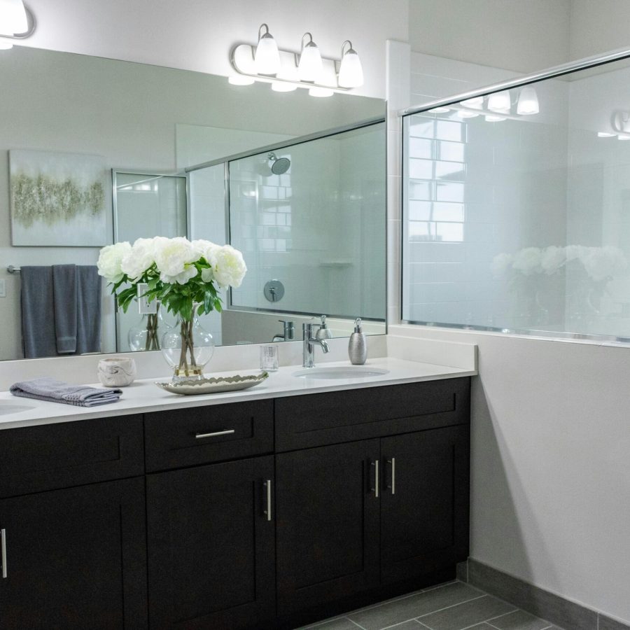 pexels-framedbydavinci-3946665 (1) Home remodeling contractor in the Upper West Side. Modern bathroom with dark wood vanity, white countertop, dual sinks, a large mirror, decorative lighting, a glass-enclosed shower, and a vase of white flowers for decor.